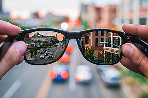 Two hands hold a pair of glasses in front of a blurry background. Through the glasses, the scenery of a city street is visible.